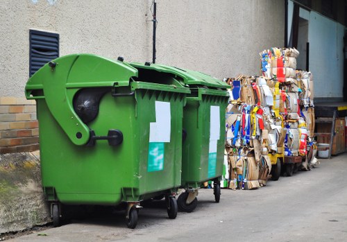 In the foreground, there are two green wheeled rubbish bins made of durable plastic with smooth surfaces, located on a paved area adjacent to a building wall. The bins are positioned side by side, with the right bin slightly behind the left one, and both have hinged lids closed on top. The wall behind them is a plain light grey concrete with a small, dark vent visible near the top left corner. To the right of the bins, there are multiple large stacks of flattened cardboard boxes and cartons piled up against the building, showing various colours and printed labels, with some cardboard pieces spilling slightly onto the ground. The ground is grey tarmac or concrete, with some minor dirt or debris near the base of the bins and stacks, indicating a typical outdoor waste disposal area. This scene reflects a dedicated rubbish collection or waste management setup, likely managed by a local waste removal service such as Business Waste Removal Finchley, operating near Finchley in London, designed for efficient waste disposal and recycling.