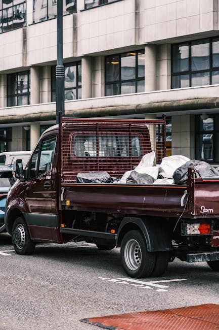 A small burgundy flatbed truck parked on a city street, filled with various types of waste materials including large white and black plastic bags, some of which appear to contain household rubbish or 